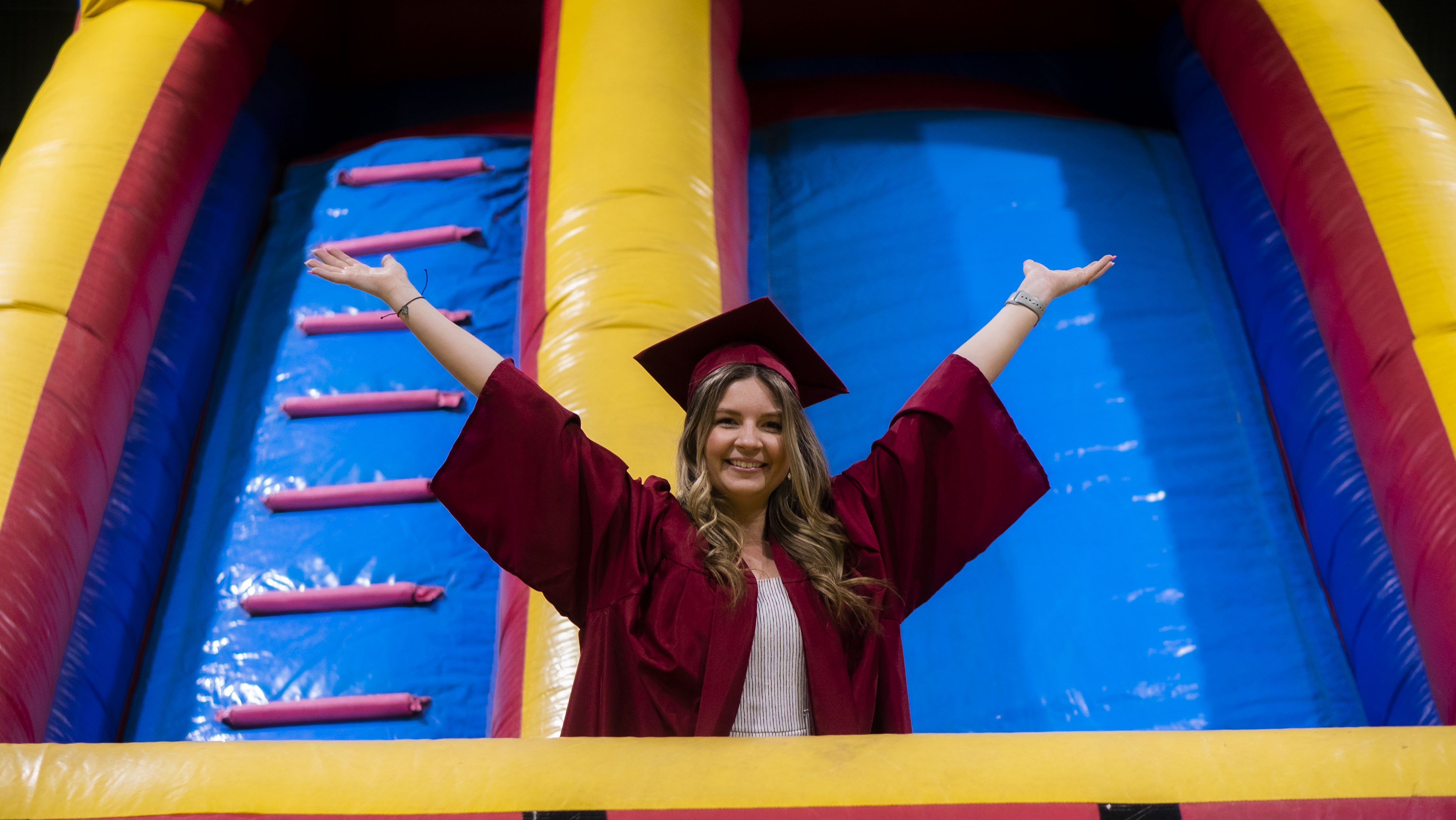 Graduate Celebrating with Inflatable Slide A joyful graduate raising her arms triumphantly in front of a colorful inflatable slide, dressed in a maroon cap and gown.