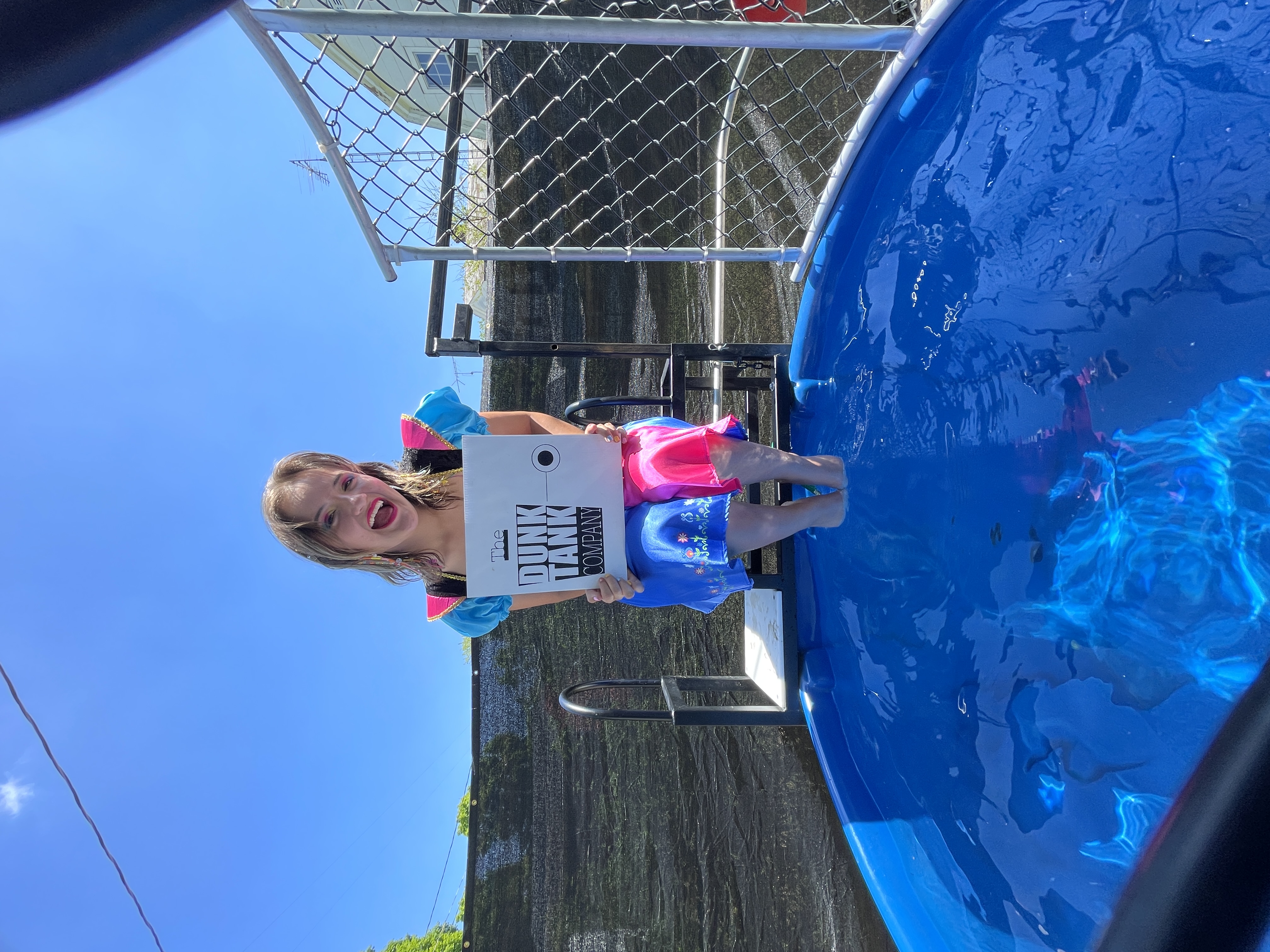 Dunk Tank Rentals by The Dunk tank Company are a great way to bring smiles to all! Girl sitting over the dunk tank holding The Dunk Tank Company Sign